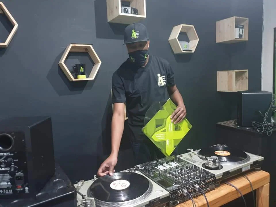 A DJ4Life instructor wearing a branded black t-shirt and cap stands behind a DJ setup in a studio. He is holding a bright green vinyl record sleeve while demonstrating mixing techniques on two Technics turntables.